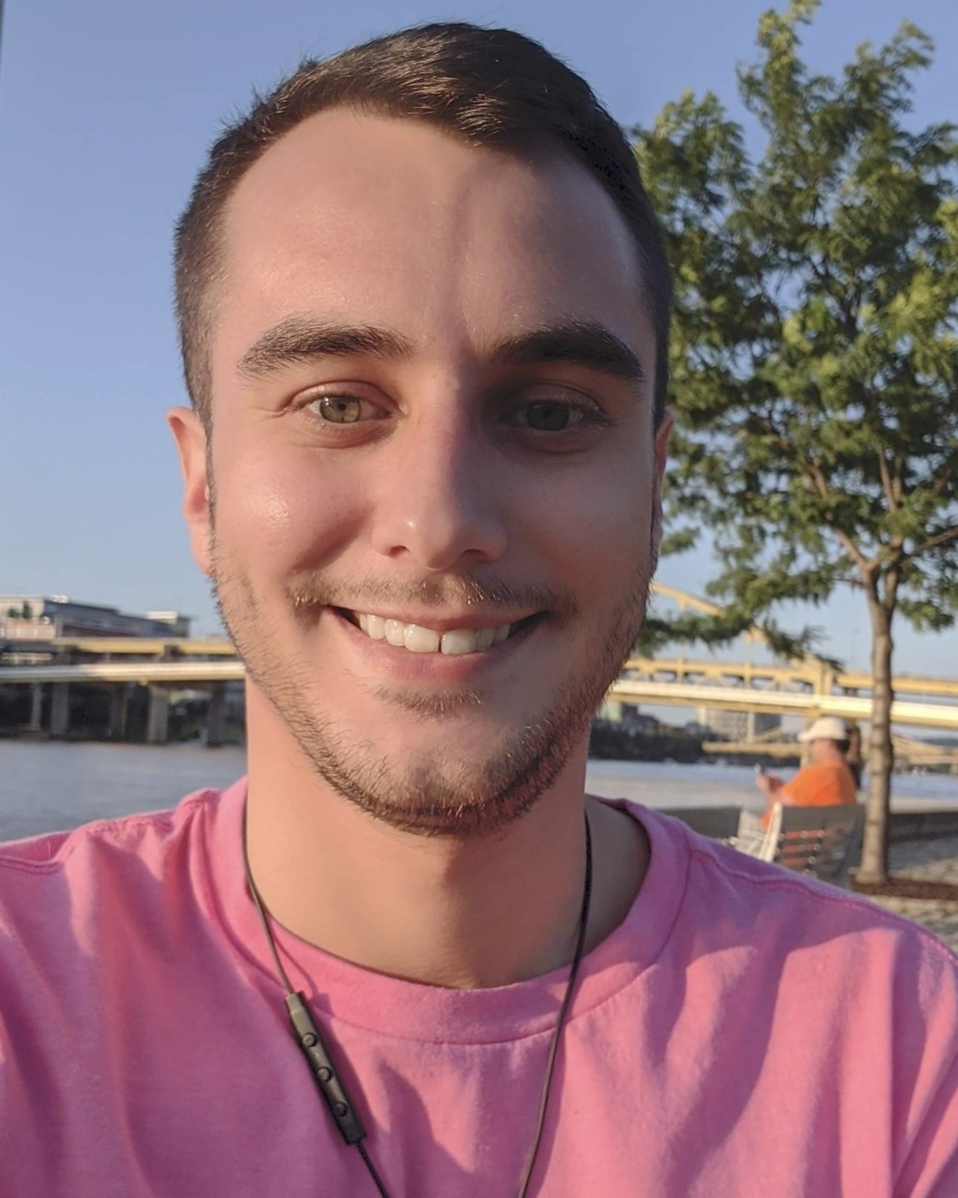 Chris outdoors in a pink shirt with trees and blue sky behind him